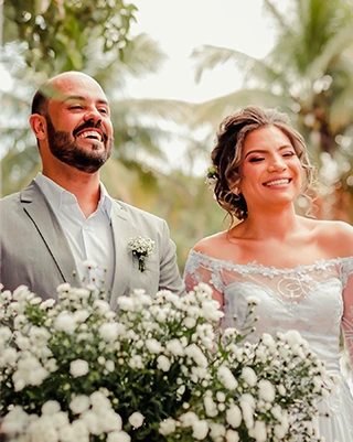 Noivos sorrindo ao ar livre, com buquê de flores brancas em primeiro plano